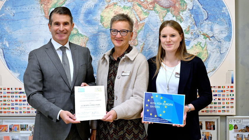 Europaminister Eric Beißwenger (links) überreicht der Einrichtungsleiterin Christl Eiler (Mitte) und der Hausleiterin Juliane Fleissner (rechts) die Europa-Urkunde für das AWO Haus für Kinder am Campeon Park in Neubiberg.